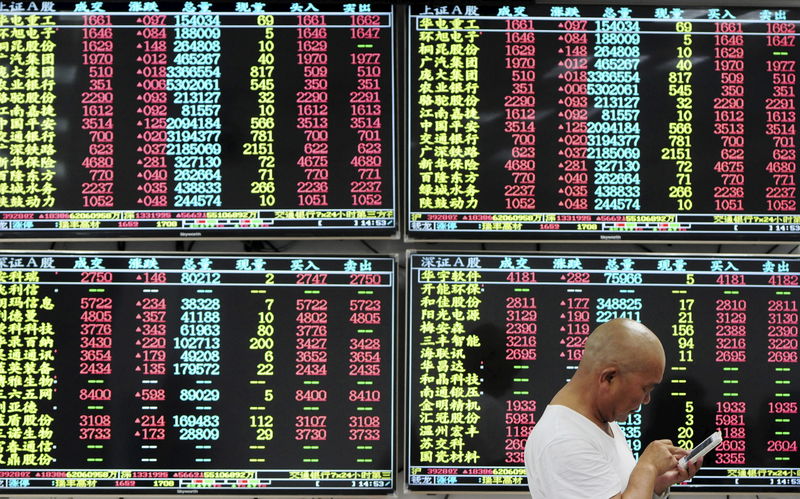 © Reuters. An investor looks at his mobile phone in front of electronic screens showing stock information at a brokerage house in Jiujiang