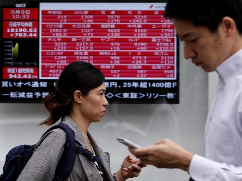 © Reuters. Pedestrians holding their mobile phone walk past an electronic board showing the various stock prices outside a brokerage in Tokyo