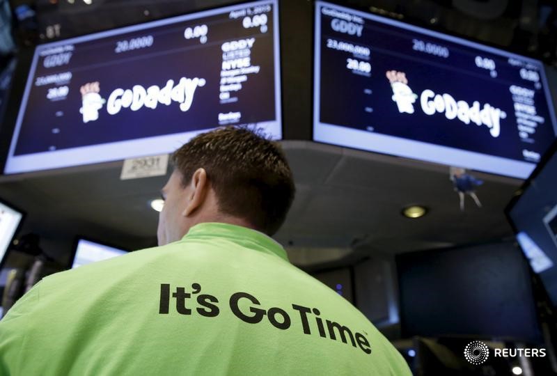 © Reuters. Man wearing a shirt featuring one of the slogans of web hosting company GoDaddy stands below screens with the company's logo, as it makes its initial public offering at the New York Stock Exchange