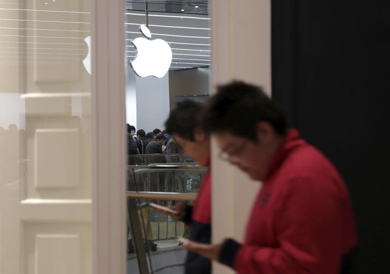 © Reuters. A logo of Apple Inc. is reflected on a mirror as a man looking at his mobile phone walks past near an Apple Store at Joy City department store in Tianjin