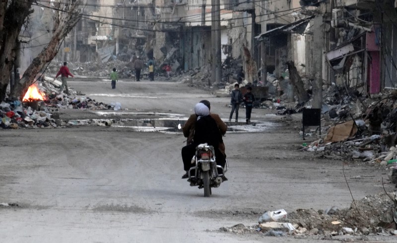 © Reuters. A man and a woman ride a motorcycle along a street filled with debris of damaged buildings in Deir al-Zor