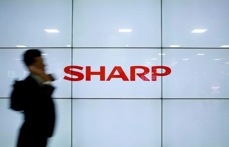 © Reuters. A man using his mobile phone walks past Sharp Corp's liquid crystal display monitors showing the company logo in Tokyo