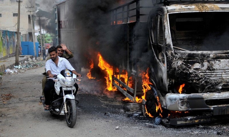 © Reuters. Men ride a motorcycle past a lorry which was set on fire by protesters in Bengaluru