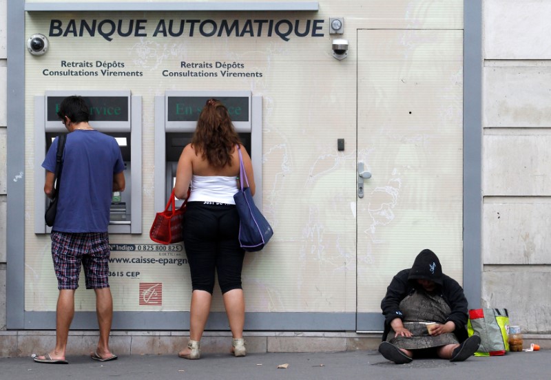 © Reuters. An homeless woman sits on a footpath while people withdraw money from ATMs along a main road in central Paris