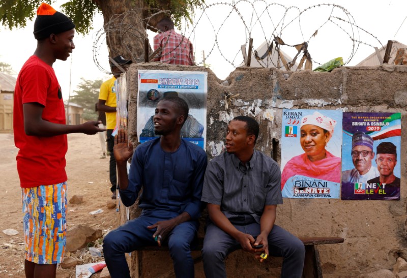 © Reuters. Youth sit around a wall pasted with election posters, after the postponement of the presidential election in Yola, Adamawa State