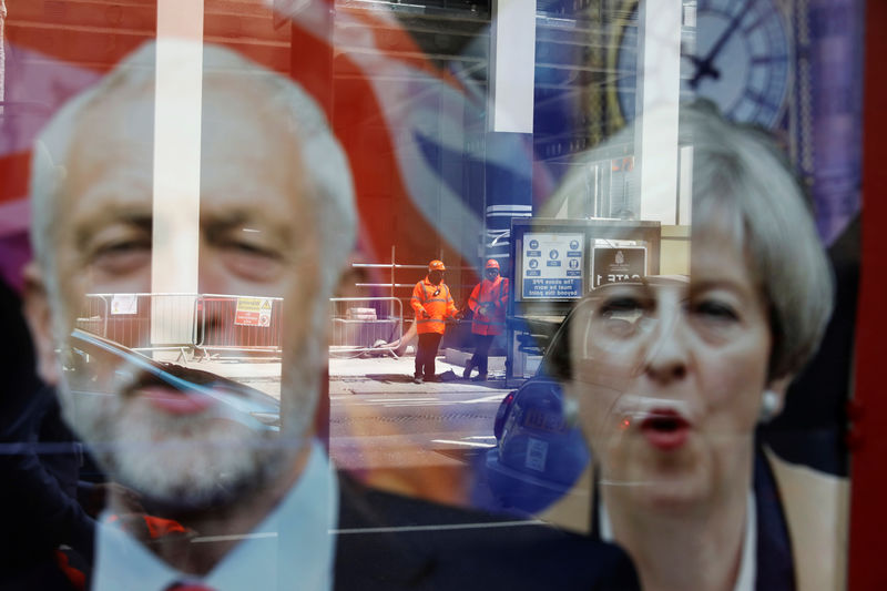 © Reuters. Los trabajadores con equipos de protección se reflejan en el escaparate de una tienda con imágenes de Theresa May y Jeremy Corbyn