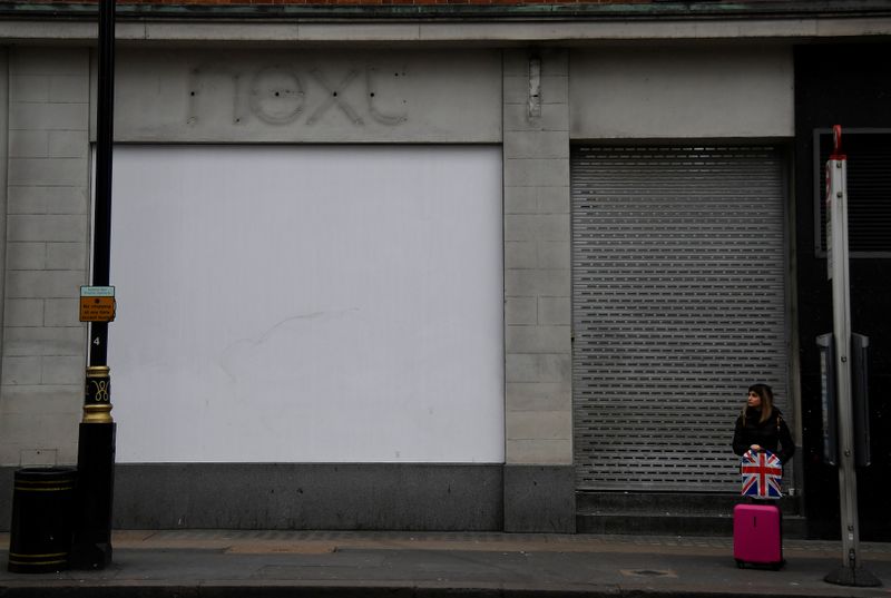 &copy; Reuters. FILE PHOTO: A shopper waits at a bus stop beside signage for a former branch of the British clothing retailer Next, off Oxford Street in London, Britain
