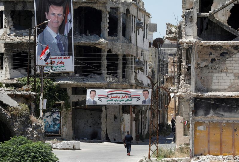 &copy; Reuters. رجل يسير بجوار  لافتات انتخابية عليها صورة الرئيس السوري بشار الأسد مبتسما على مبنى كتهدم تعرض للقصف في مدينة حمص في سوريا يوم 23 مايو أيار 2021